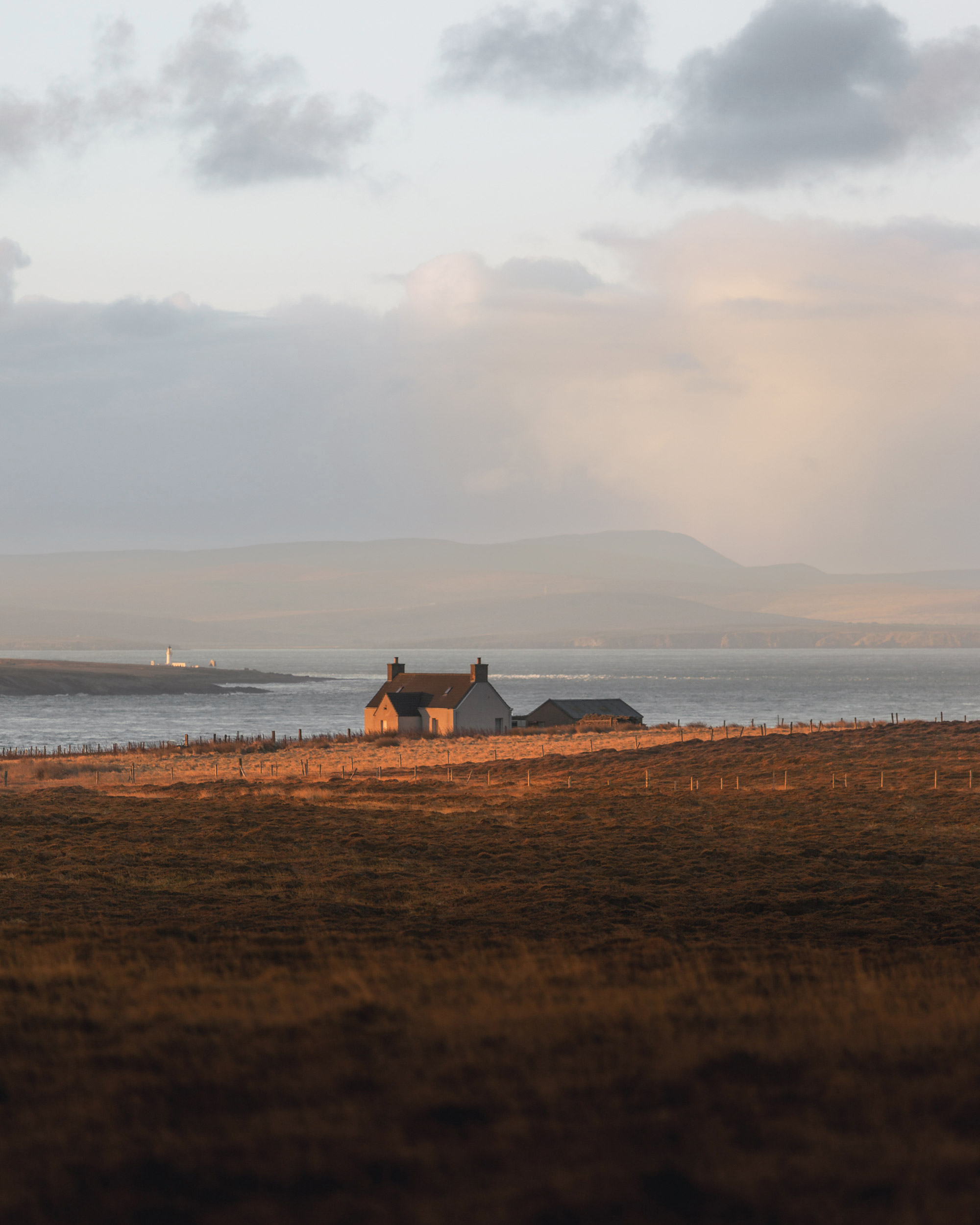 The image features a house situated in a field next to a body of water. The landscape showcases elements of nature, including grass and the shore along the beach. Above, the sky is adorned with clouds, hinting at a beautiful sunset on the horizon.
