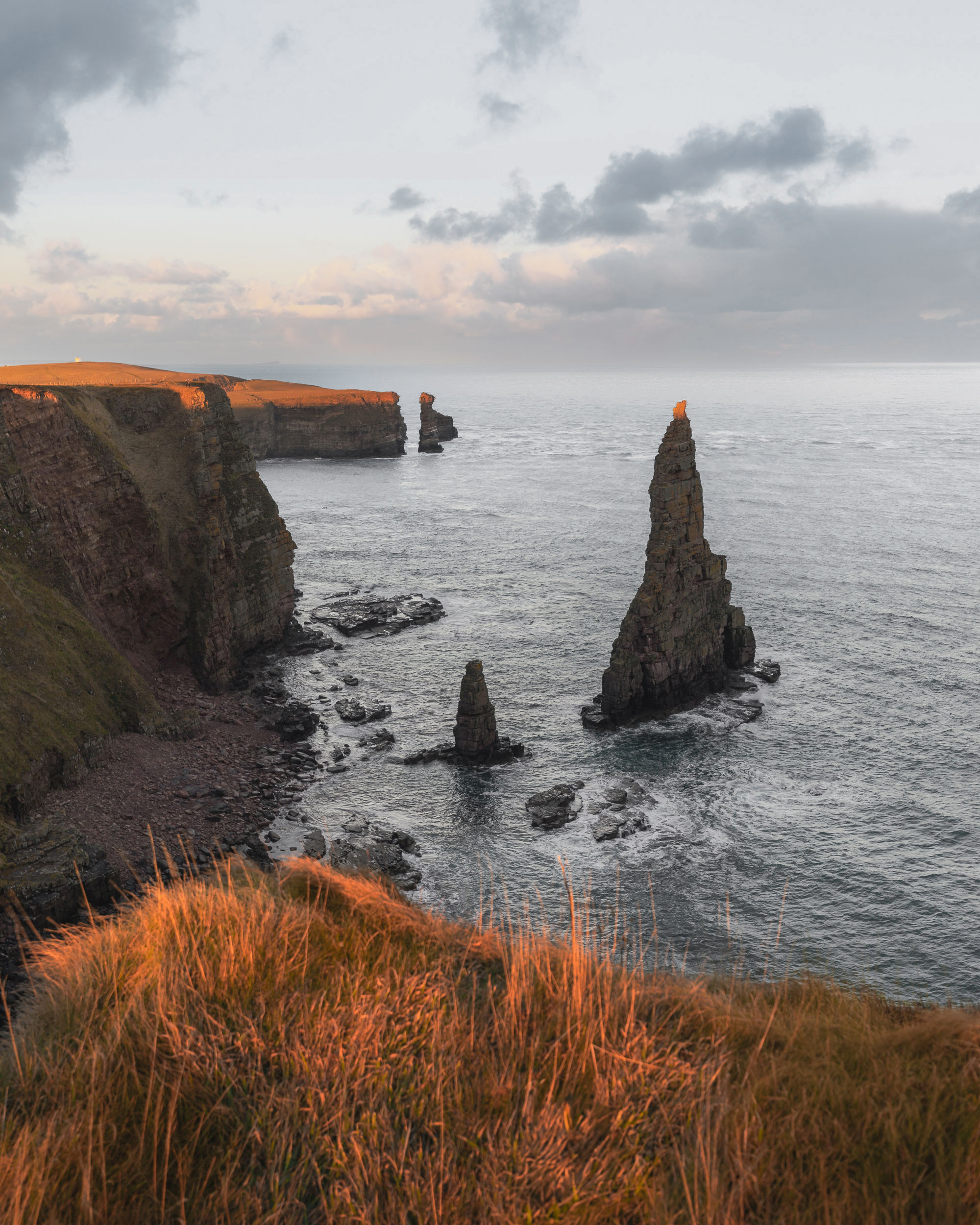 The image depicts rocky cliffs alongside a body of water, likely an ocean or sea. The landscape features natural coastal formations, including headlands and promontories. The scene captures the outdoor beauty of nature, with elements such as grass and a stunning sunset in the background.