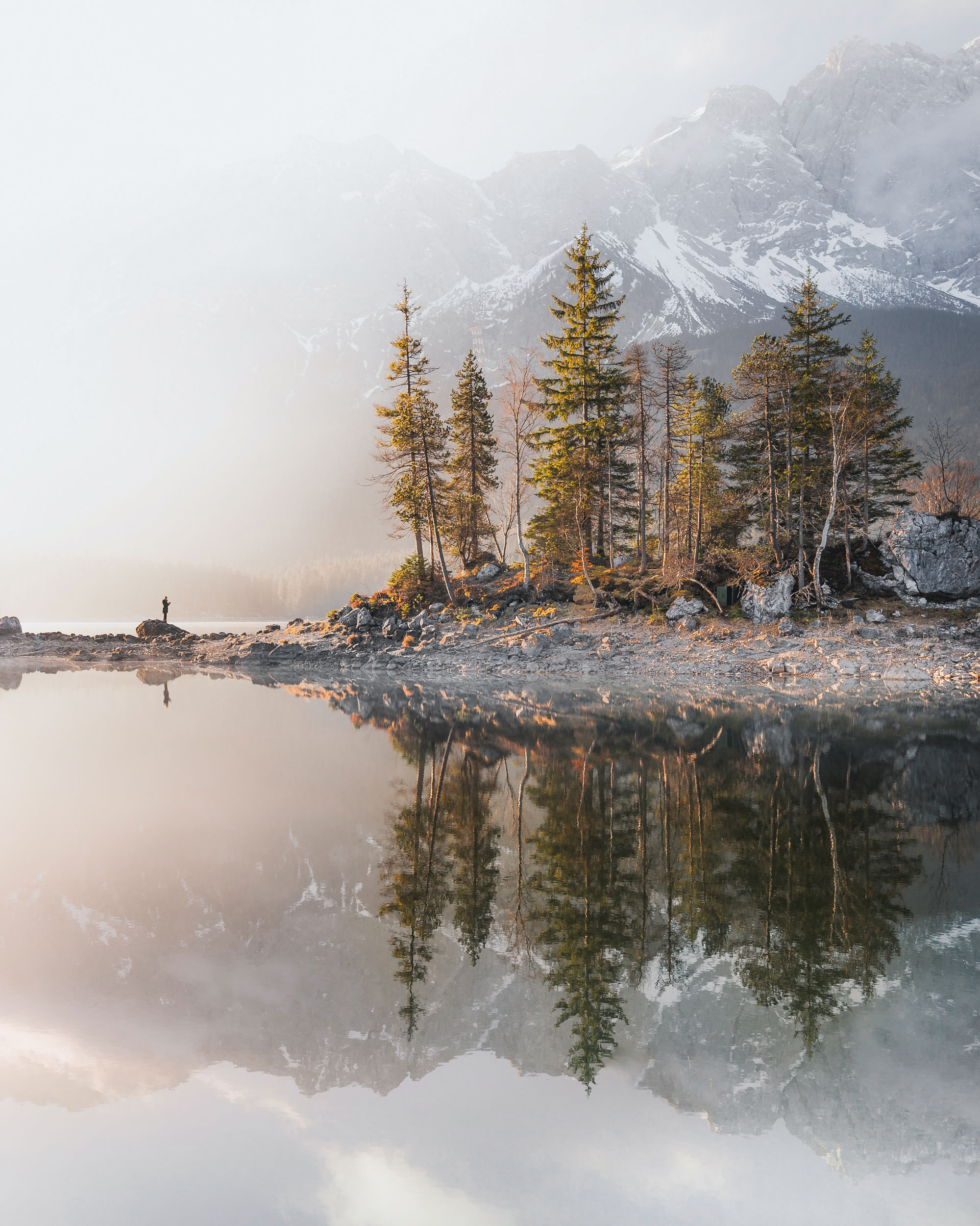 The image depicts a tranquil body of water surrounded by trees, capturing the essence of a peaceful natural landscape. The scene features reflections on the water's surface, with elements such as fog and mist adding to the serene atmosphere.