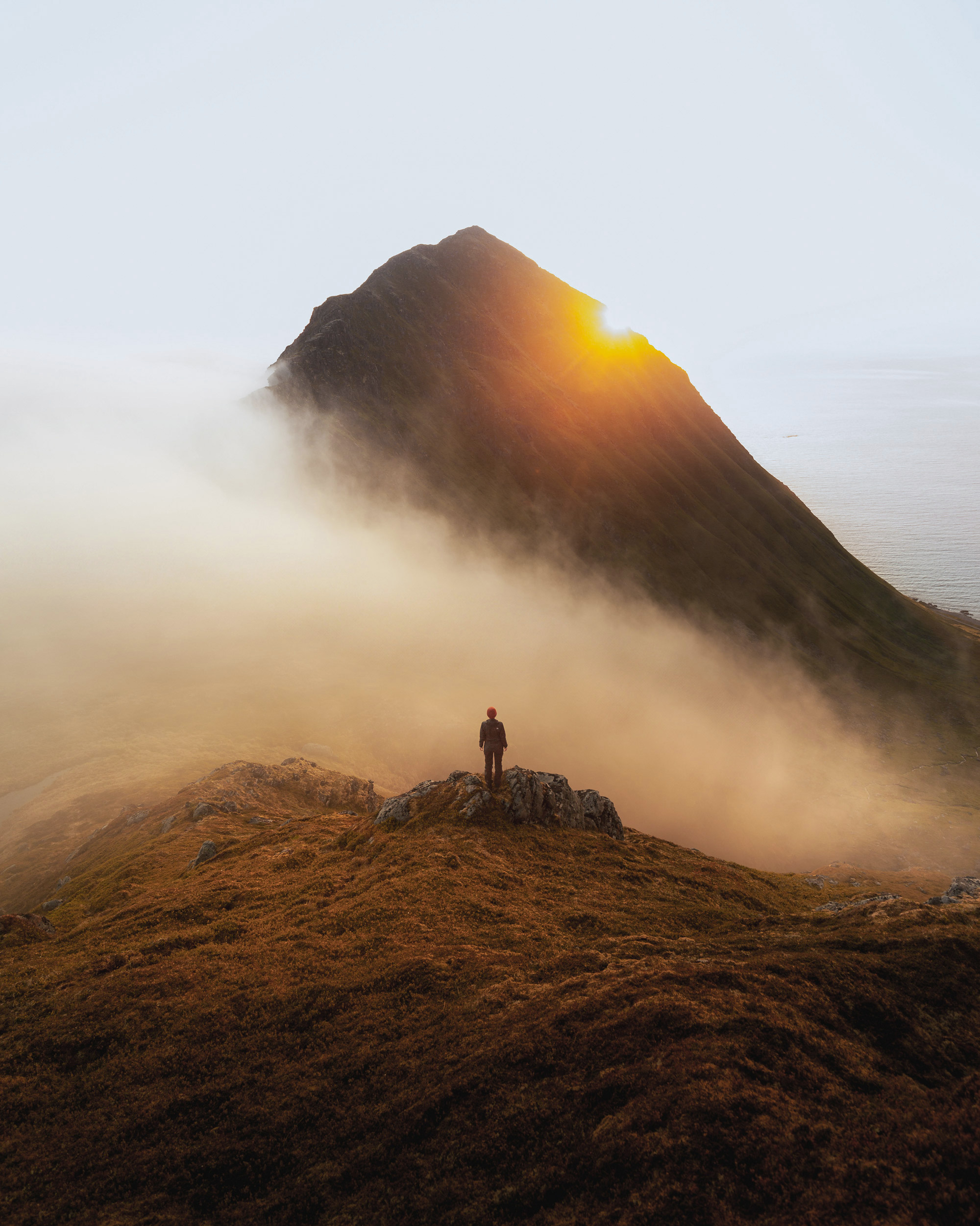 A hiker stands on a summit, overlooking a valley veiled in morning mist.  Golden light bathes the scene.