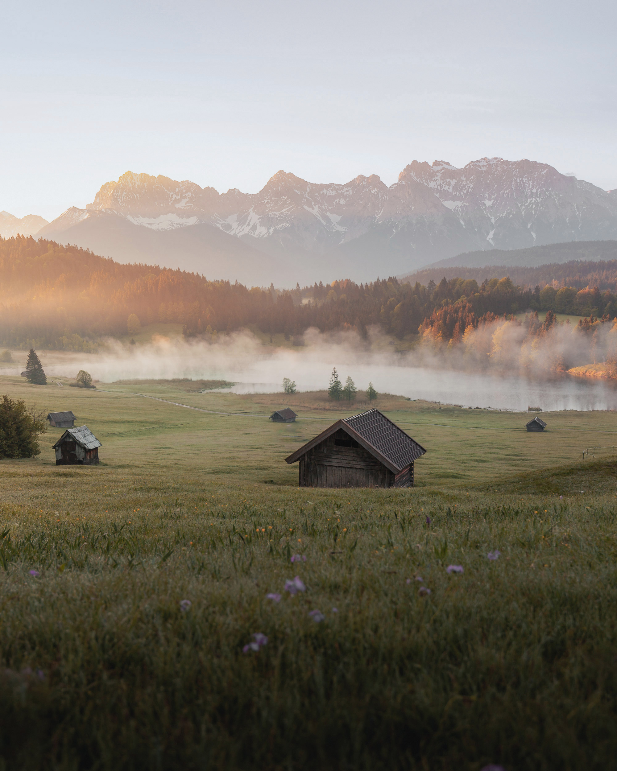 The image captures a tranquil landscape bathed in the soft light of dawn. In the foreground, a lone wooden cabin stands on a gently sloping hill, its slanted roof suggesting a rustic charm.