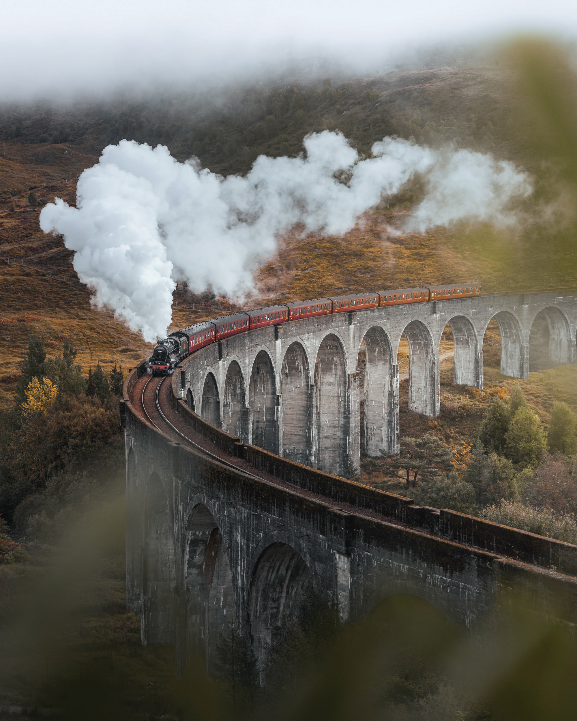 The image captures a picturesque scene of a vintage steam locomotive chugging along a serpentine viaduct that spans a lush, rolling landscape.