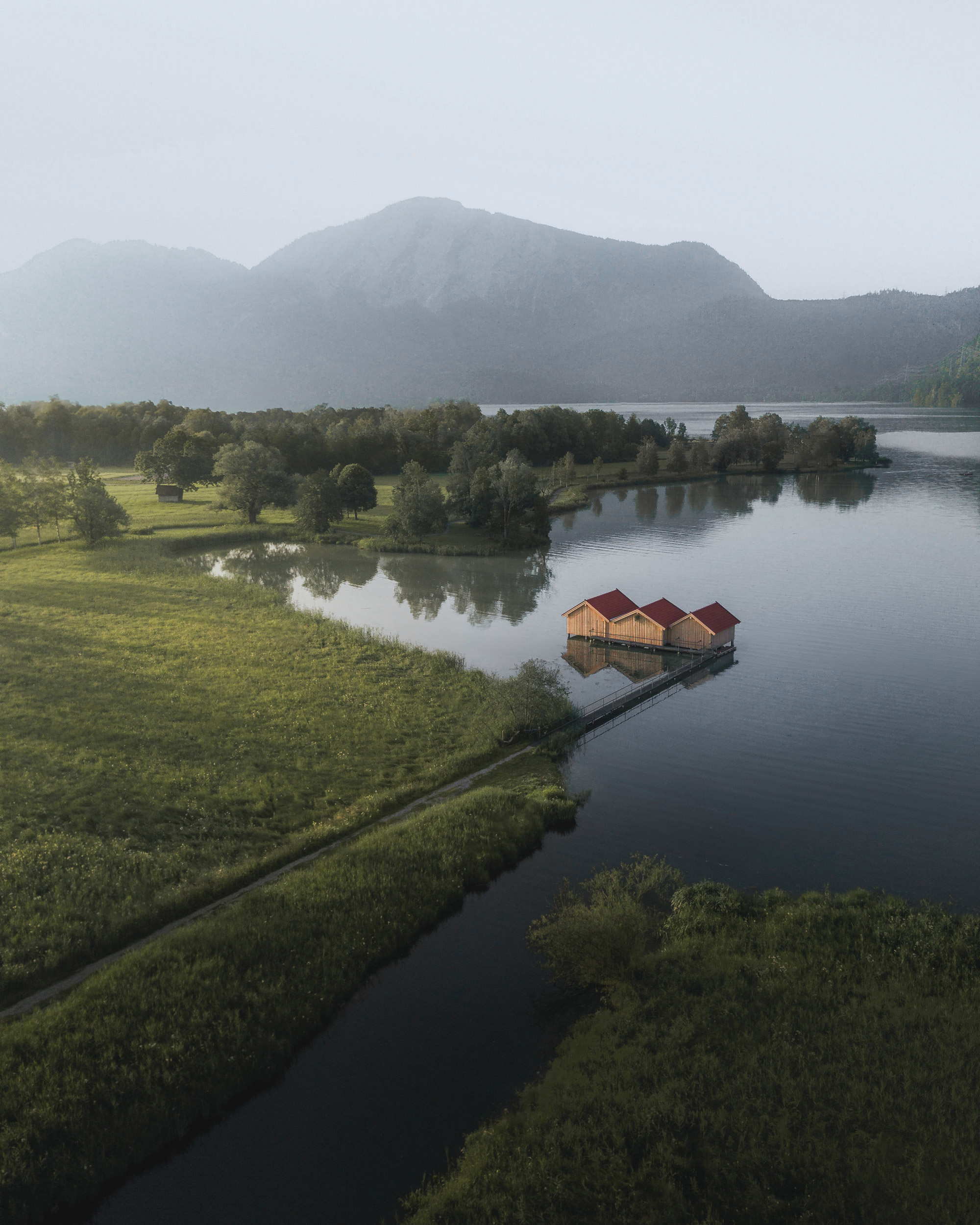 The image features a picturesque house situated by a serene lake, surrounded by natural beauty. The landscape includes lush grass and trees, with water reflecting the sky. Elements like mountains and fog in the background add to the tranquil atmosphere, while a boat is visible on the lake, suggesting recreational use of this scenic outdoor space.