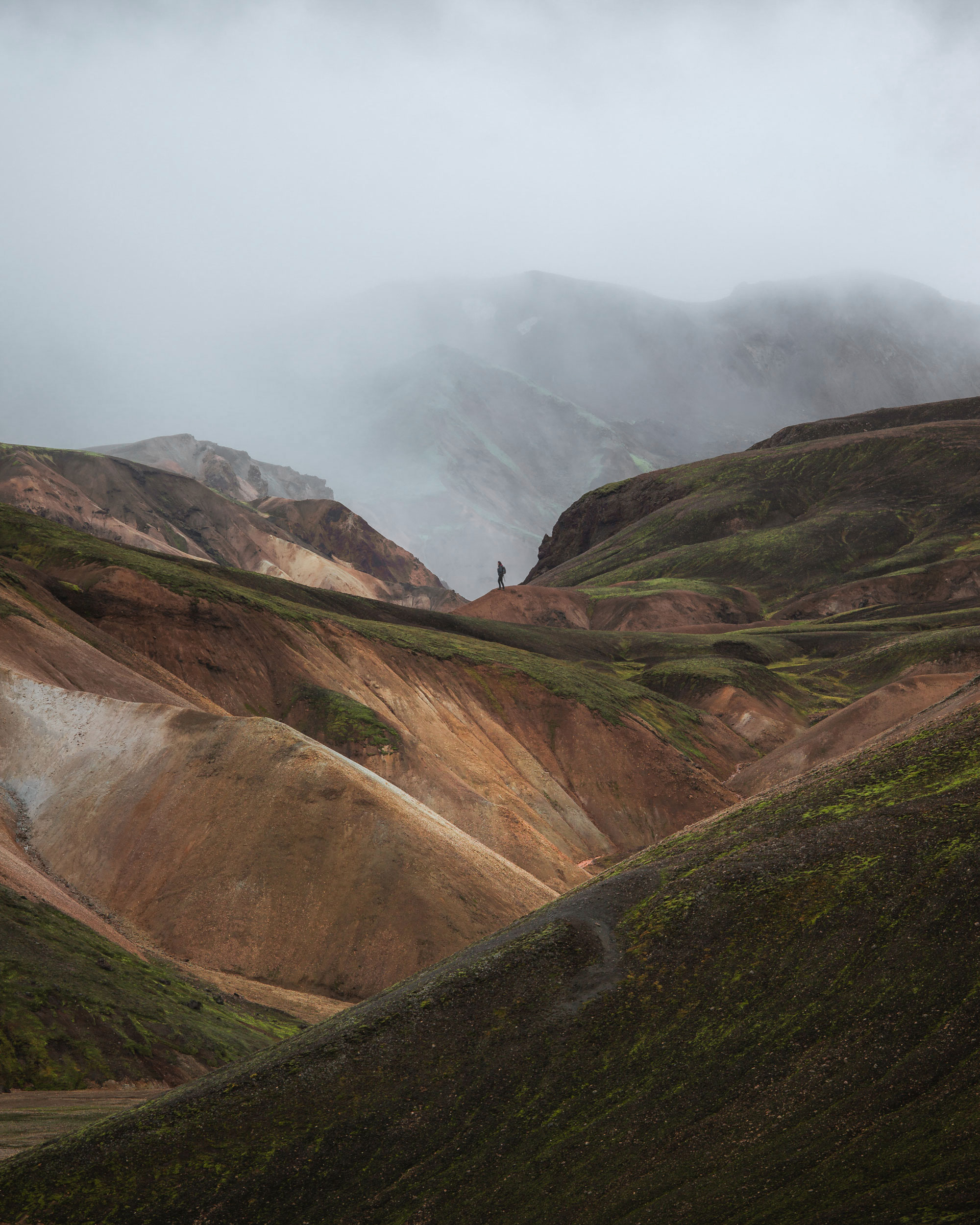 A person is standing on a mountain, surrounded by a breathtaking landscape. The scene features dynamic terrain with grass and rocky surfaces, set against a backdrop of clouds and fog that add to the natural beauty. This highland area includes valleys and hills from the mountain range, showcasing the rugged geology of the region.