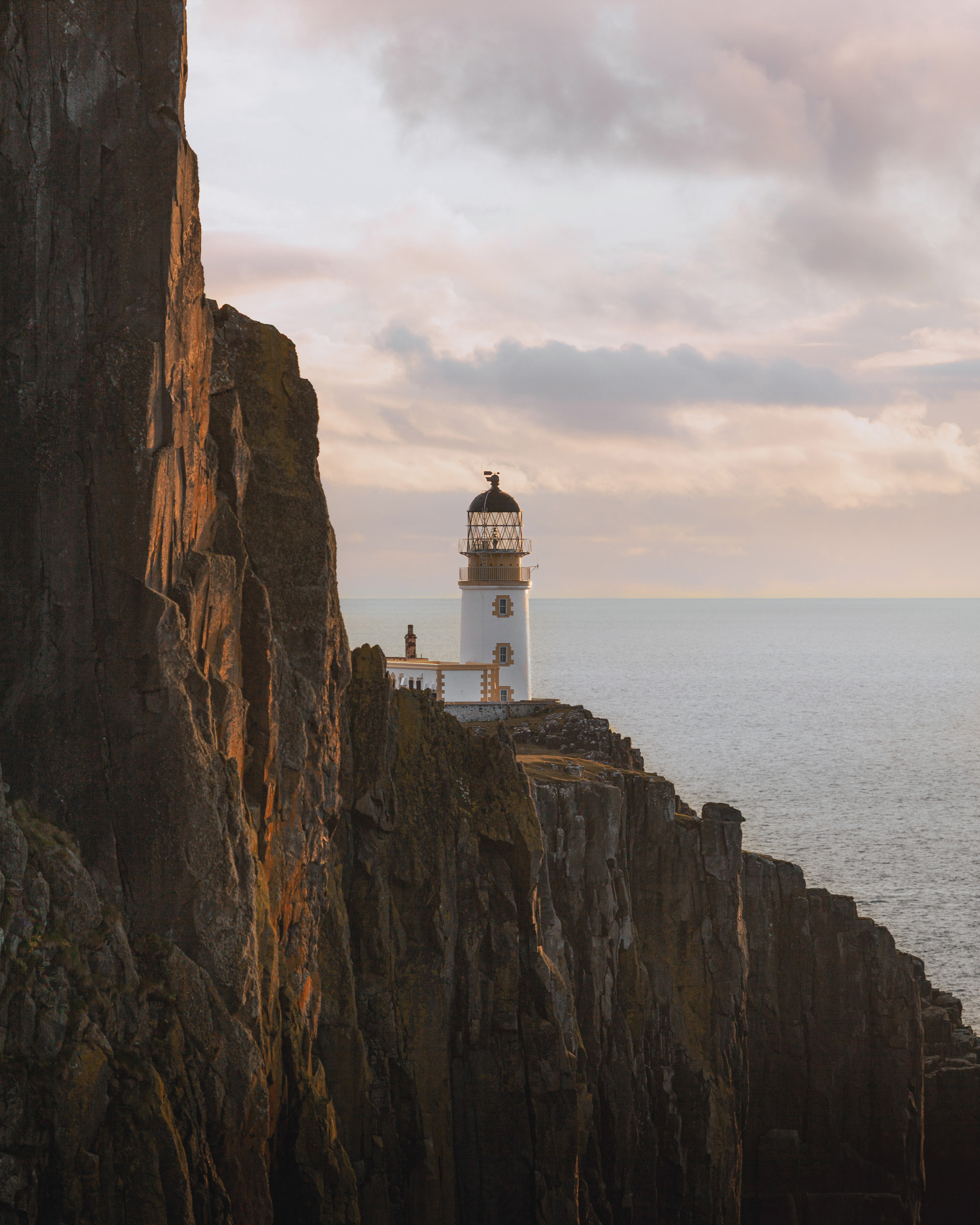 The image features a lighthouse positioned on a cliff, overlooking the sea. The structure serves as a beacon, guiding ships safely through the waters. The backdrop includes a clear sky with clouds, and the rugged landscape of rocks enhances the scenic view. This outdoor setting beautifully captures the essence of coastal life.