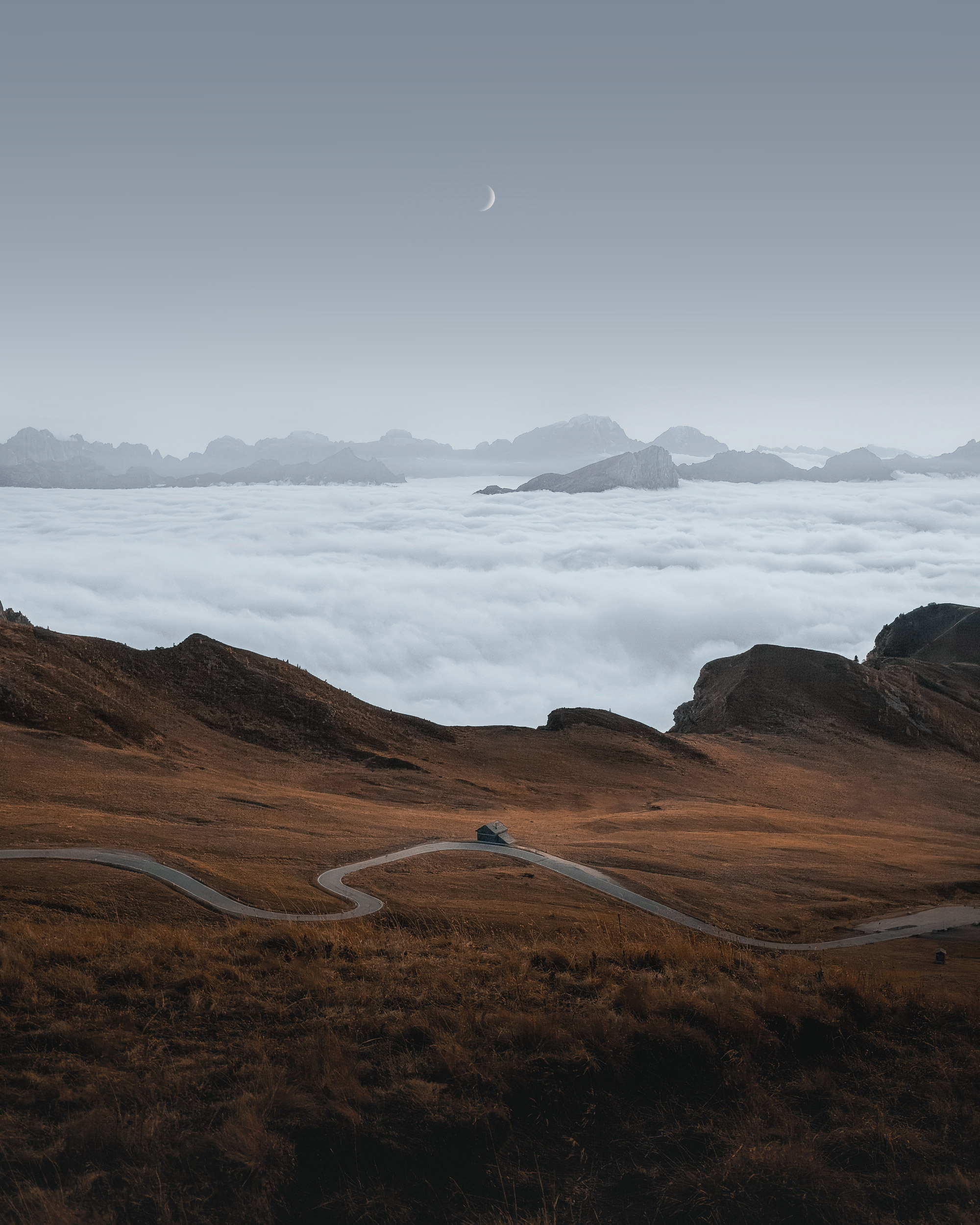 A winding mountain road above a sea of clouds with a crescent moon in the sky.