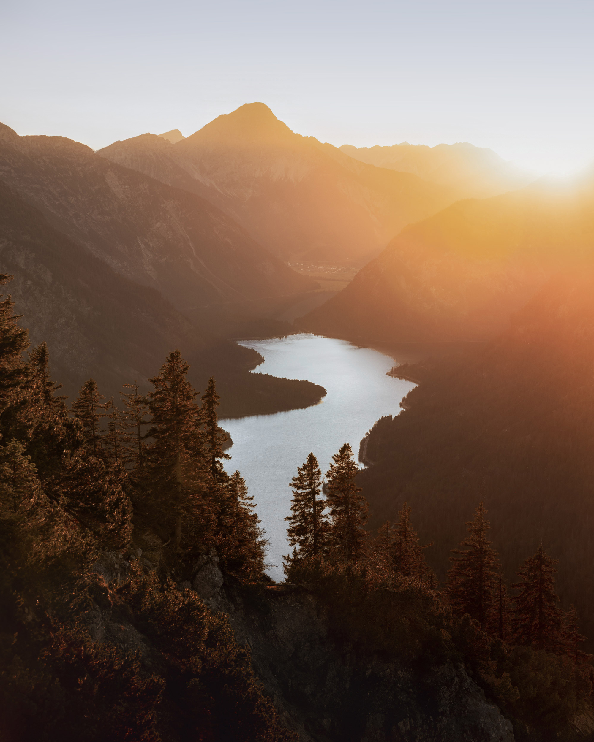The image depicts a river flowing through a serene valley, surrounded by majestic mountains and lush trees. The landscape is enhanced by the presence of a lake and enveloped in soft fog, capturing the essence of wilderness. The scene is beautifully lit by the colors of sunrise or sunset, creating a peaceful atmosphere in nature's highland setting.