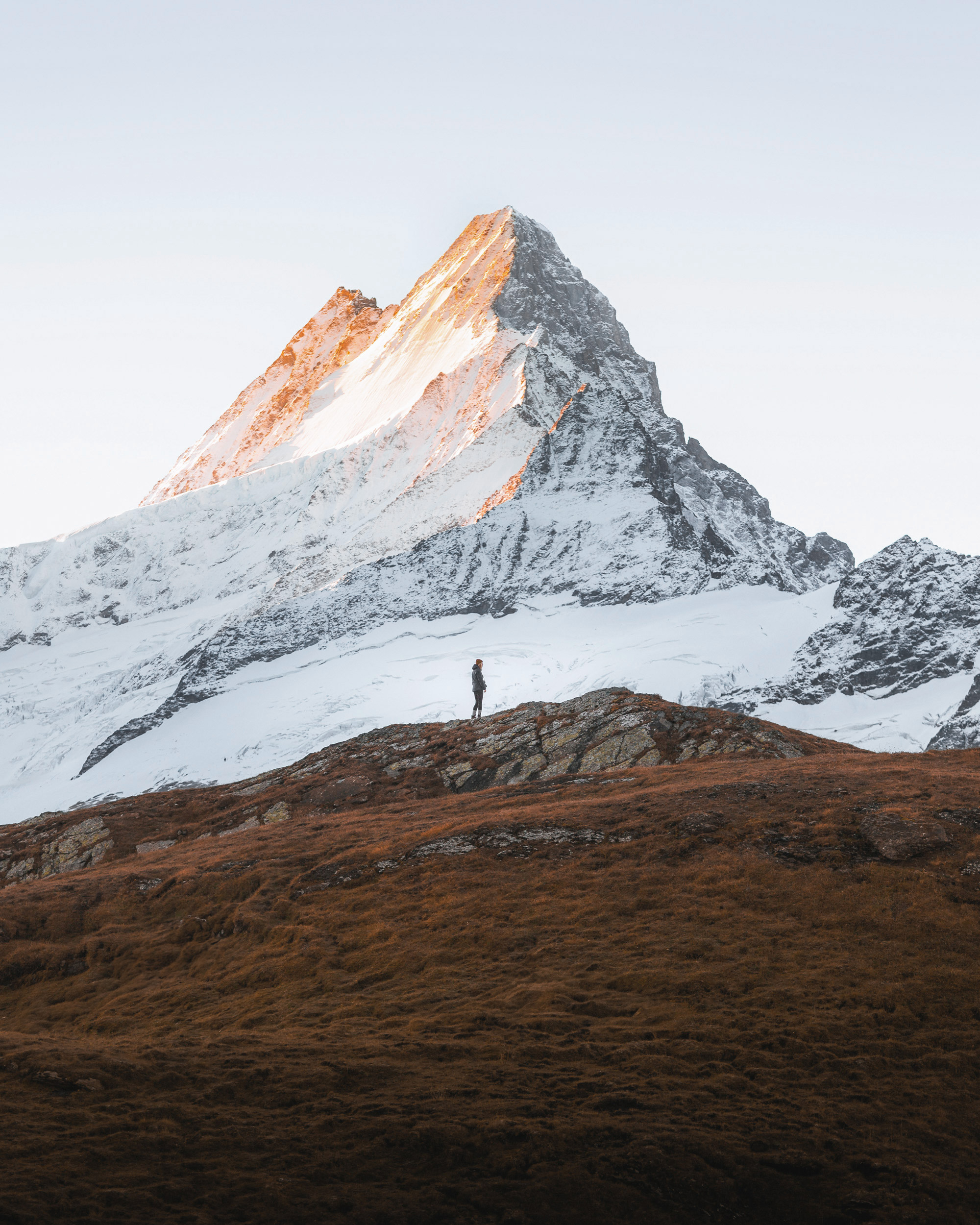 A person is standing on a mountain, enjoying the scenic views from the summit. The landscape features a beautiful mountain range with snow-covered slopes and glacial landforms