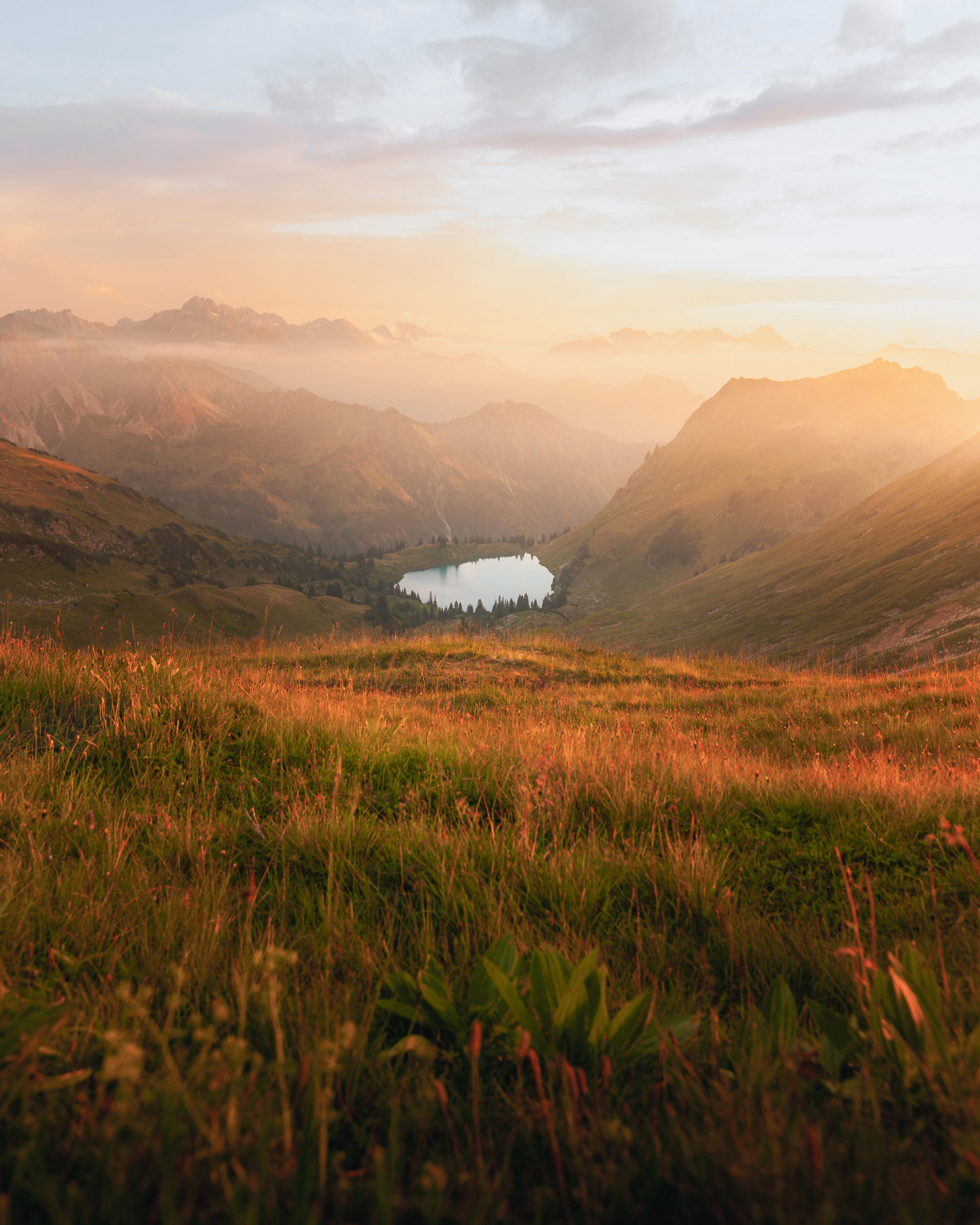 The image depicts a serene field of grass with a body of water visible in the distance. It showcases the beauty of nature, featuring elements like clouds, mountains, and a vast sky above.