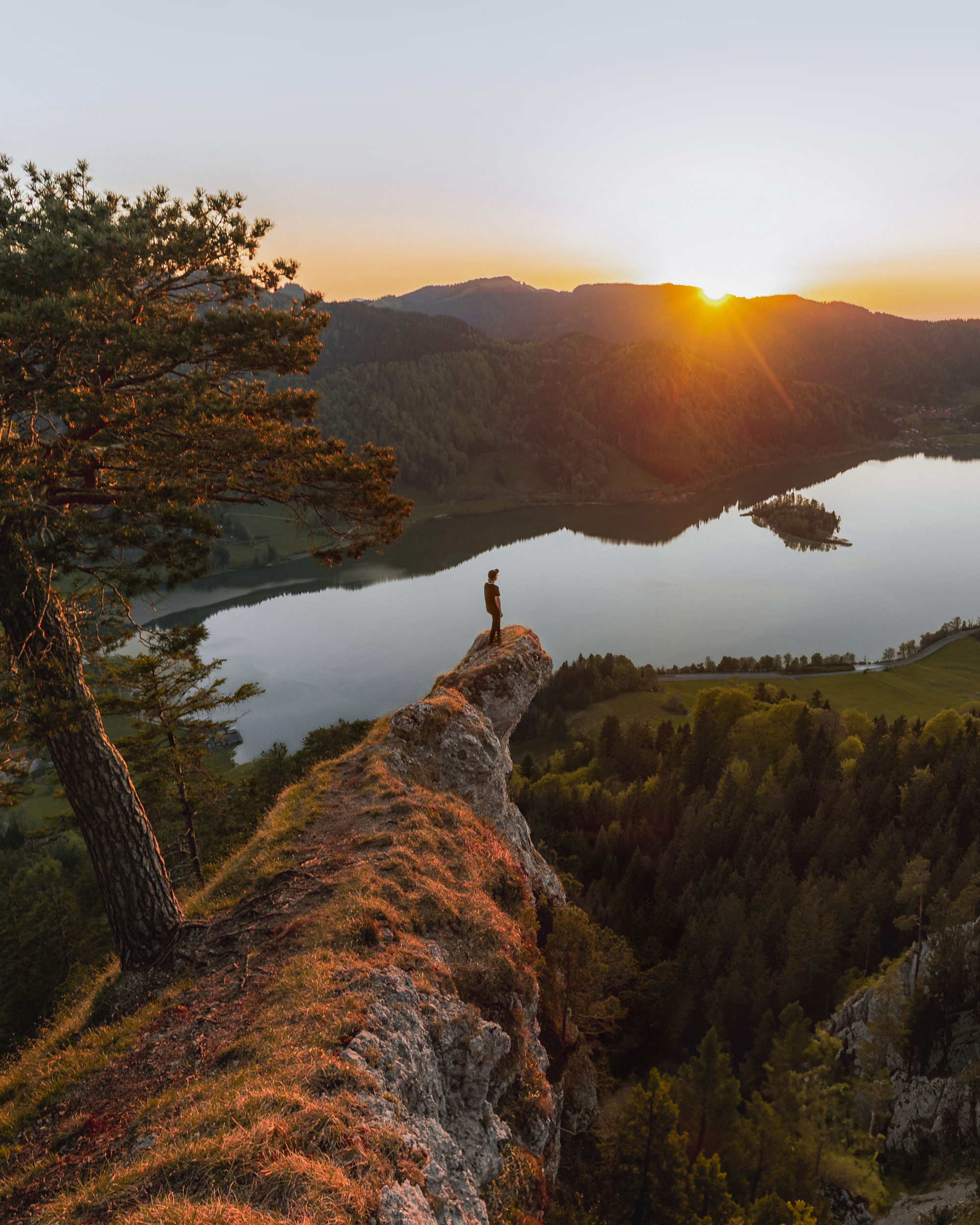 A person is standing on the edge of a cliff, overlooking a serene lake surrounded by majestic mountains. The scene captures the beauty of the outdoors during sunrise or sunset, highlighting vibrant colors in the sky and reflecting off the water. Lush trees and autumn foliage enhance the natural landscape, creating a picturesque wilderness setting.