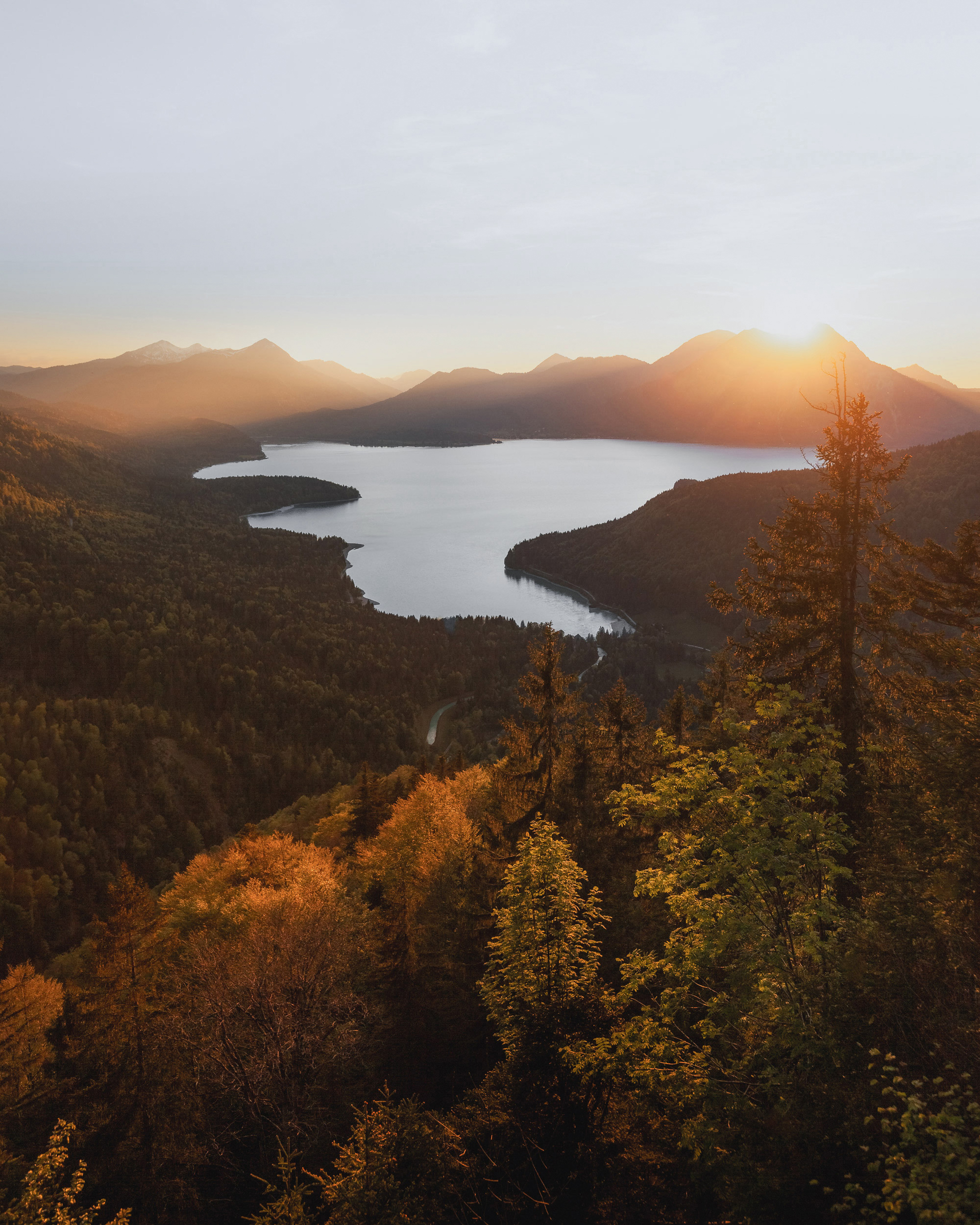 A large, calm lake is situated in a valley surrounded by towering mountains. The lake's surface appears smooth and reflective, mirroring the colors of the sky. The water's color transitions from a light, almost turquoise hue in the shadowed areas to a lighter grayish-blue in areas illuminated by the setting sun.