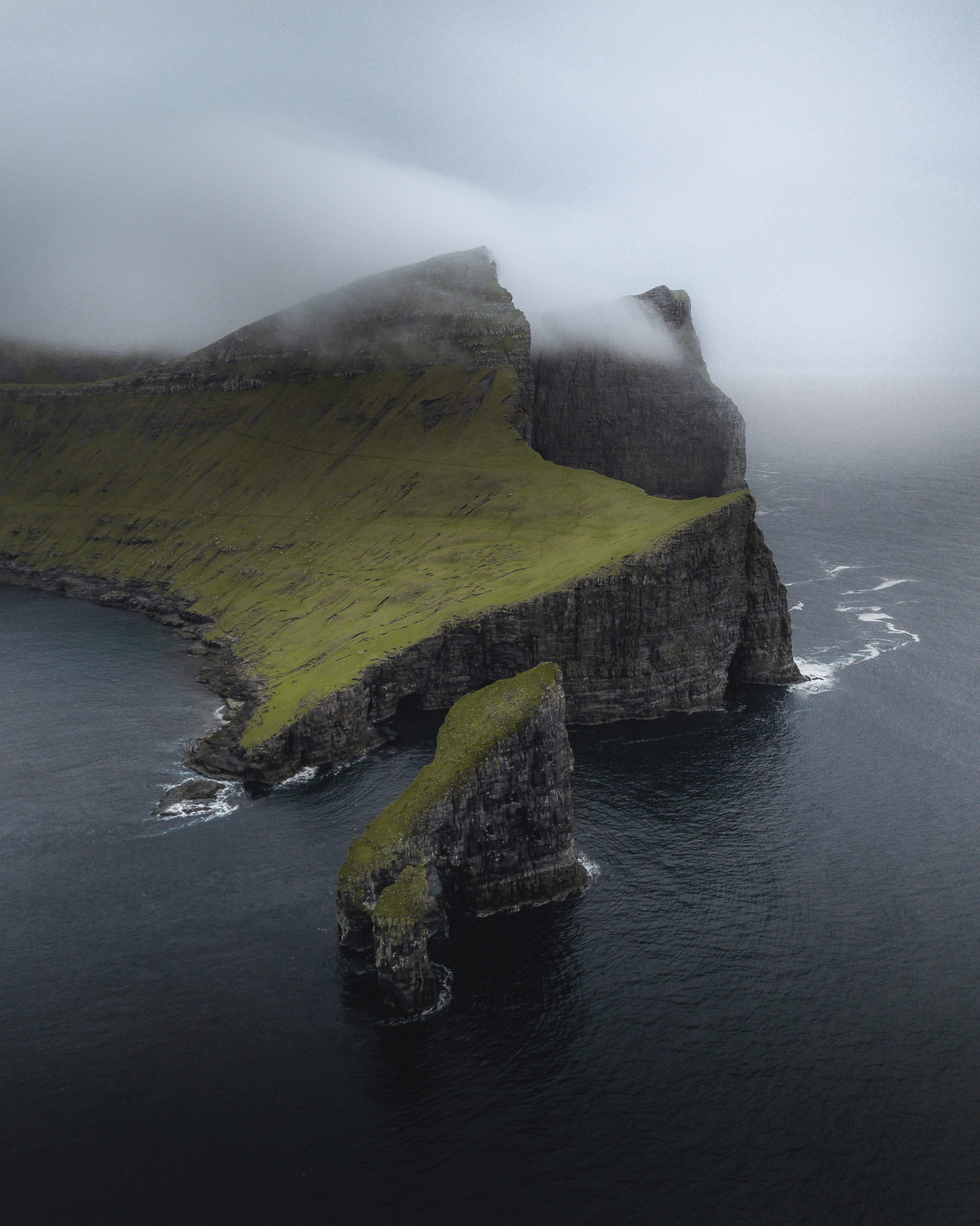 Aerial view of Drangarnir sea stacks and the towering sea cliffs of Vágar island, Faroe Islands, shrouded in low-hanging mist.