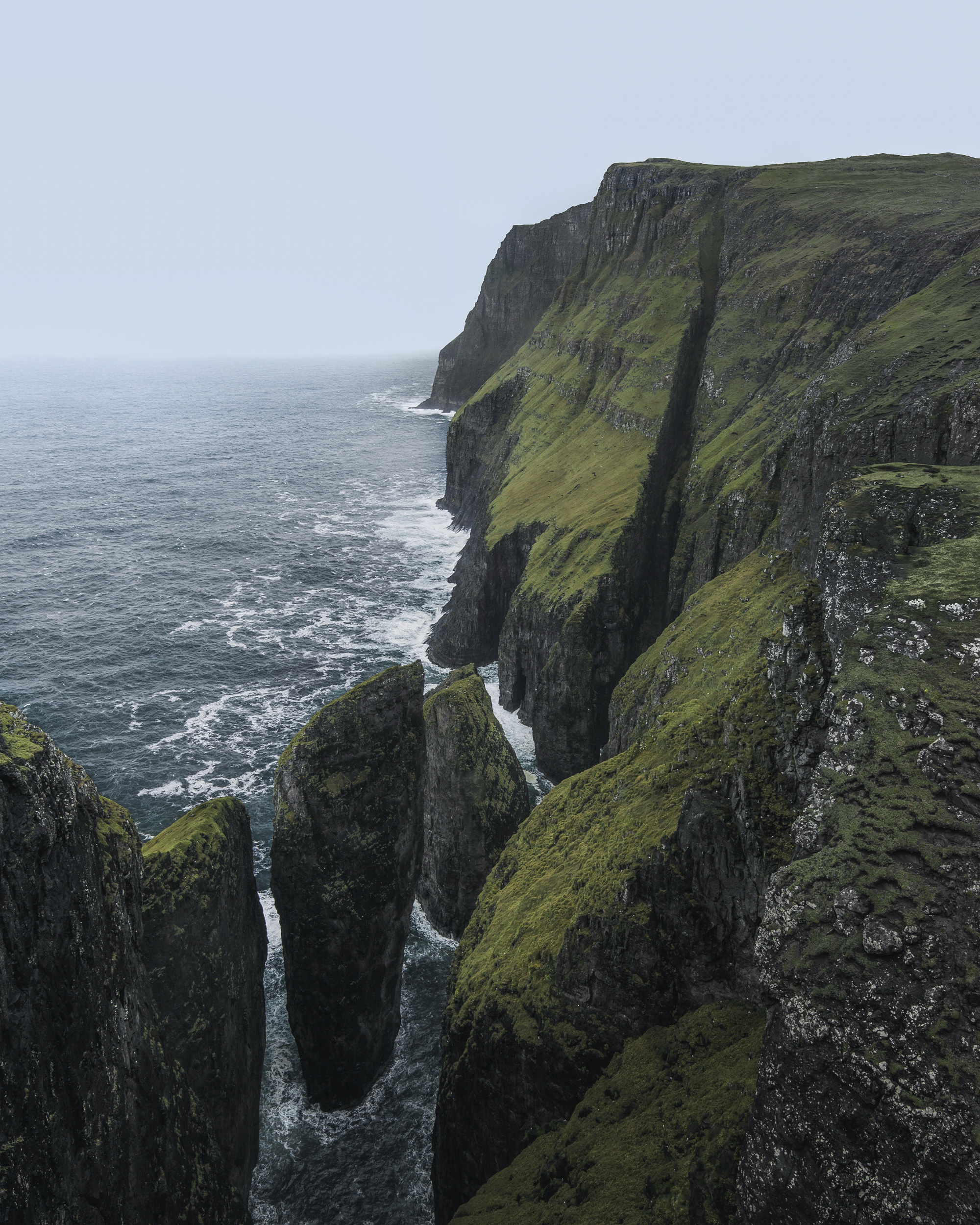 The imposing vertical rock formations of Dunnesdrangar rise from the churning North Atlantic waters beneath overcast skies in the Faroes.