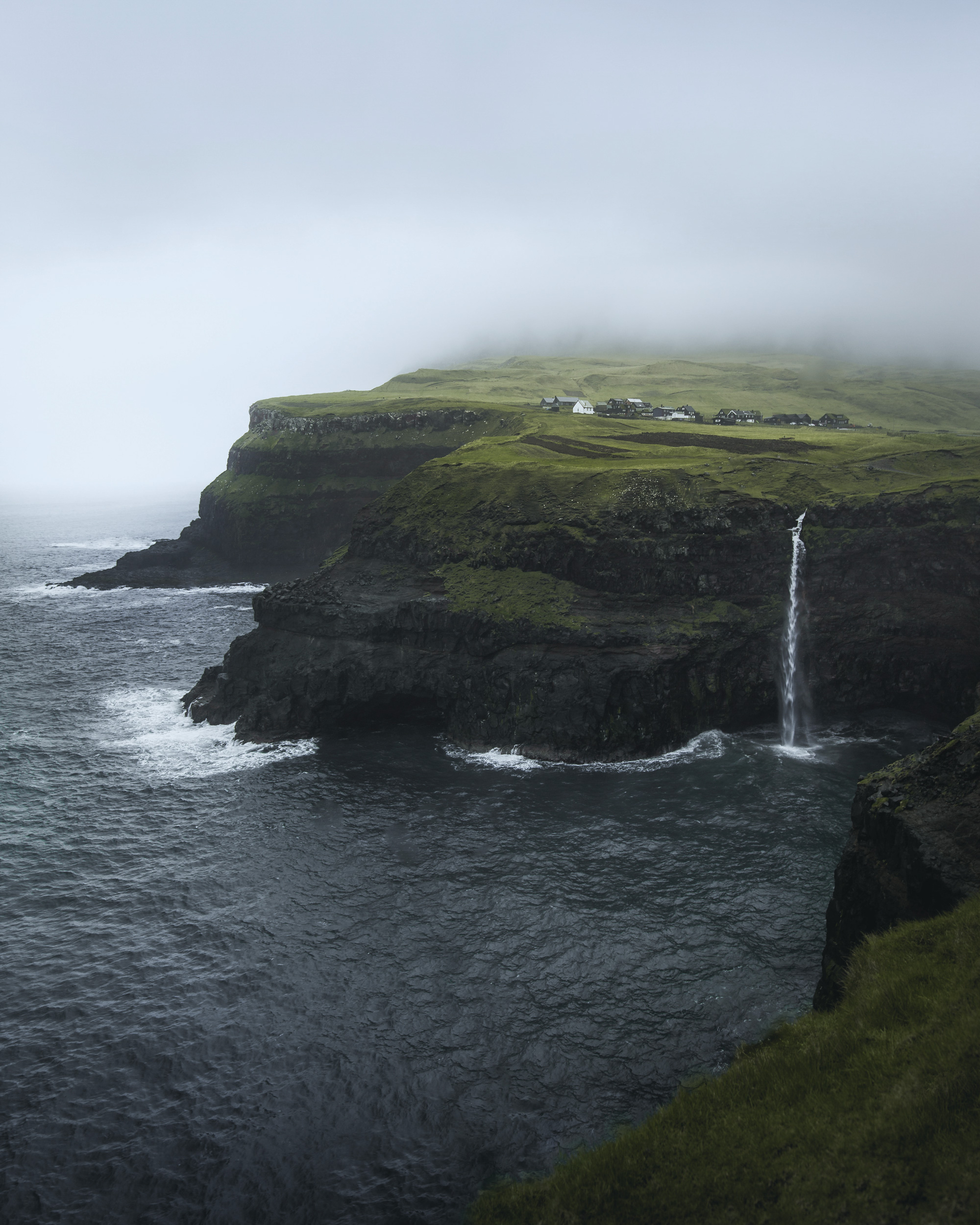 The image depicts a stunning waterfall cascading over a cliff and into a body of water below. Surrounded by the natural beauty of the Faroe Islands, the scene includes elements like mountains, cliffs, and coastal landforms. The atmosphere appears serene, possibly enhanced by fog in the background, highlighting the rugged landscape of Gasadalur.