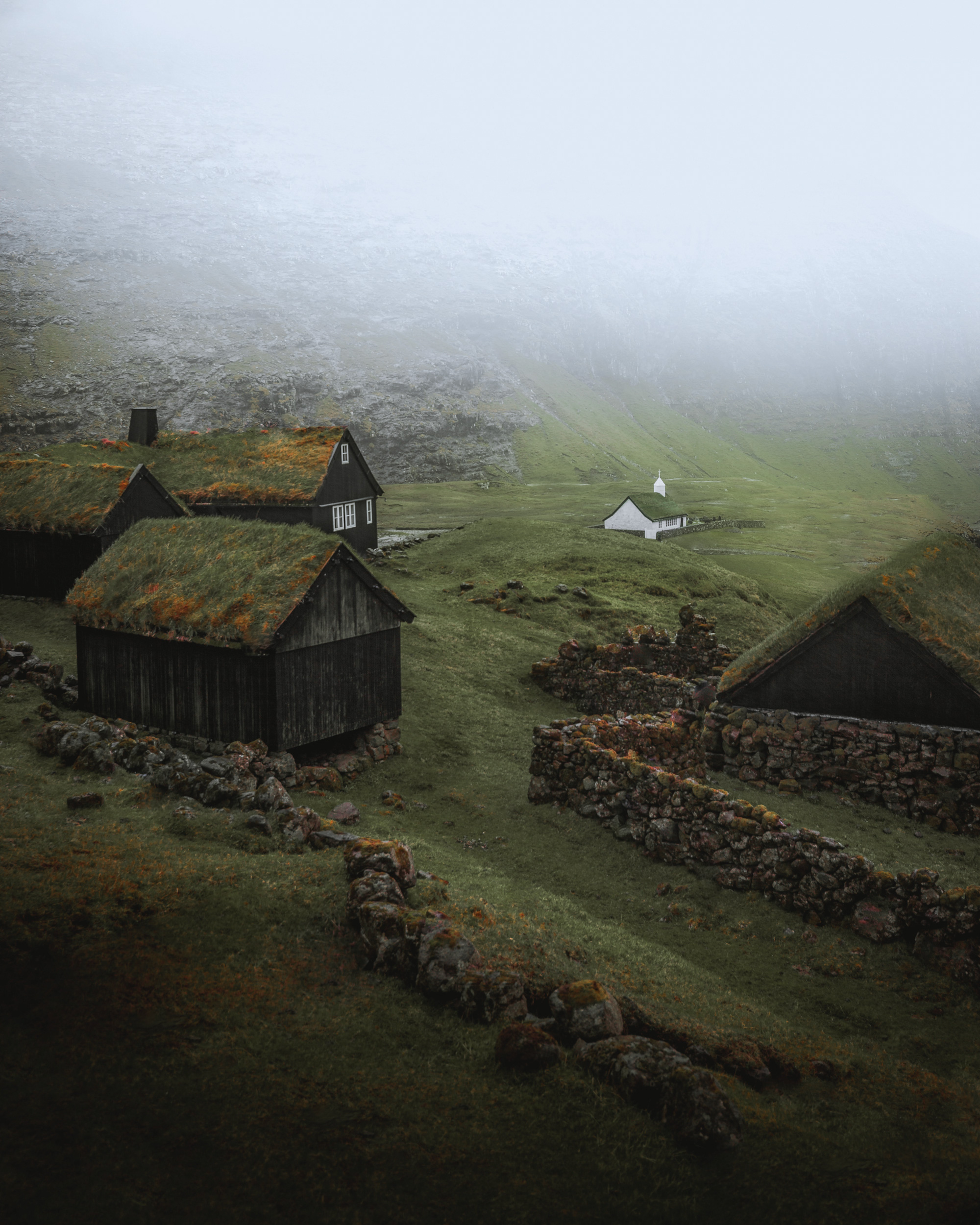 The image depicts a group of houses situated on a hill in Saksun, Faroe Islands. The landscape features lush grass and is enveloped in a serene fog, with clouds lingering overhead. This picturesque scene illustrates the natural beauty of the mountainous terrain and tranquil environment.