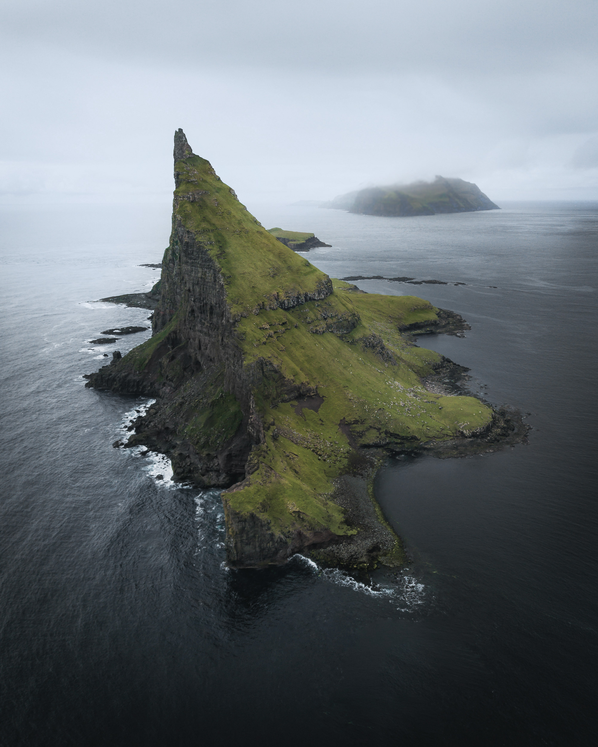 The dramatic, grassy slopes and sheer basalt cliffs of Tindholmur under a moody, overcast sky, typical of the Faroese weather.